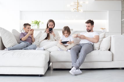 Mother, father, and two elementary school-aged children on couch in luxury home, using connected smartphones and tablet computer. 